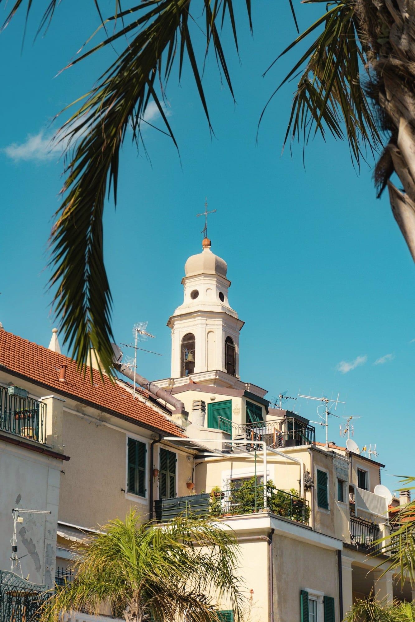 Un campanile tra le palme e le case dalle persiane verdi, a Pietra Ligure